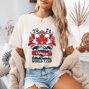 Peut inclure: T-shirt cr&egrave;me avec un motif graphique pour la Coupe du Monde Canada 2026. Le motif comprend une feuille d'&eacute;rable rouge, le texte "CANADA WORLD CUP" et les chiffres "20 26". Le t-shirt est port&eacute; par une personne.