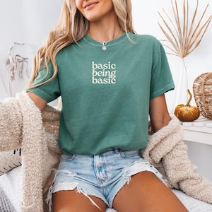 Puede incluir: Una camiseta verde azulado con las palabras "basic being basic" en blanco. La camiseta se combina con pantalones cortos de mezclilla y una rebeca color crema. En el fondo hay calabazas decorativas y una cesta de calabaza tejida.