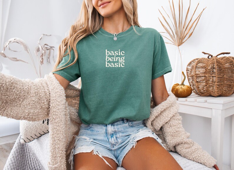 Puede incluir: Una camiseta verde azulado con las palabras "basic being basic" en blanco. La camiseta se combina con pantalones cortos de mezclilla y una rebeca color crema. En el fondo hay calabazas decorativas y una cesta de calabaza tejida.
