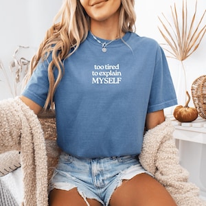 May include: A blue short-sleeved t-shirt featuring the white text "too tired to explain MYSELF" printed on the chest. The shirt has a crew neckline and a relaxed fit, paired with light blue denim shorts and a textured beige cardigan draped over.