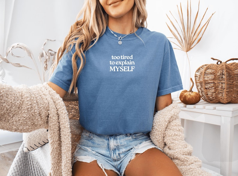 May include: A blue short-sleeved t-shirt featuring the white text "too tired to explain MYSELF" printed on the chest. The shirt has a crew neckline and a relaxed fit, paired with light blue denim shorts and a textured beige cardigan draped over.