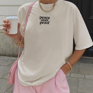 May include: An off-white short-sleeve t-shirt featuring the black text "peace over proof" printed on the chest. The shirt is paired with pink and white pinstriped shorts and a light pink shoulder bag, accessorized with a gold chain necklace and bracelets.