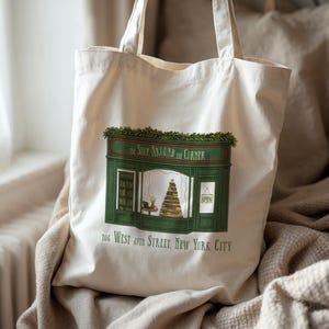 May include: Cream-colored tote bag featuring a green and red illustration of a shop facade with the text "The Shop Around the Corner" and "106 West 69th Street, New York City." The shop has a Christmas tree in the window.