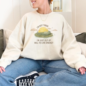 Może przedstawiać: Kremowa bluza z napisem "I'M NOT TIRED I'M JUST OUT OF WILL TO LIVE ENERGY" oraz grafiką żaby na liściu lilii wodnej ze słońcem. Bluza jest zestawiona z niebieskimi dżinsami i czarnymi trampkami.