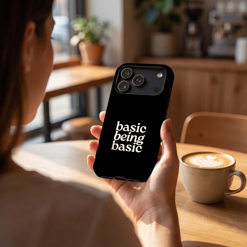 Black Basic Being Basic iPhone case with white text, held in hand at a warm coffee shop table with a latte cup in the background.