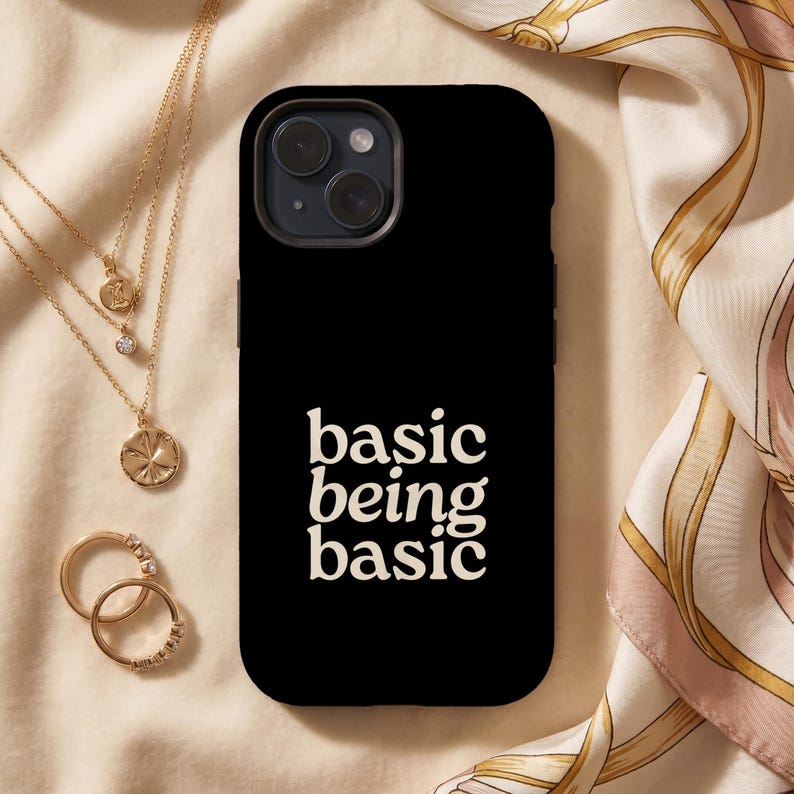 Black iPhone case with ‘basic being basic’ white text, styled on cream satin fabric with gold jewelry