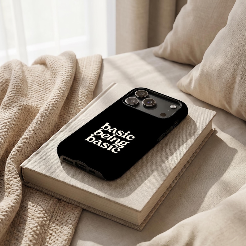Black Basic Being Basic iPhone case with white text, resting on a book in a cozy neutral-toned bed setting with knit blanket and pillows.