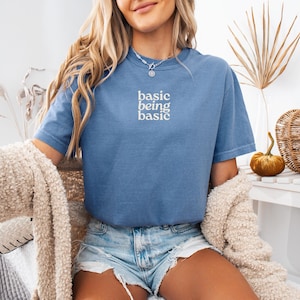Puede incluir: Una camiseta azul con las palabras "basic being basic" en escritura blanca. La camiseta se combina con pantalones cortos de mezclilla. Un su&eacute;ter beige est&aacute; drapeado sobre los hombros. Una peque&ntilde;a calabaza y una cesta decorativa est&aacute;n sobre una mesa blanca.