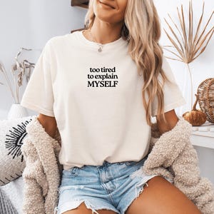 May include: A cream-colored short-sleeve t-shirt featuring the black text "too tired to explain MYSELF" printed on the center of the chest. The shirt has a crew neckline and a relaxed fit, paired with light blue denim shorts and a textured cardigan.