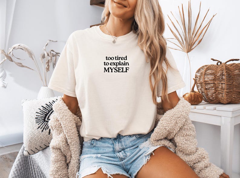 May include: A cream-colored short-sleeve t-shirt featuring the black text "too tired to explain MYSELF" printed on the center of the chest. The shirt has a crew neckline and a relaxed fit, paired with light blue denim shorts and a textured cardigan.