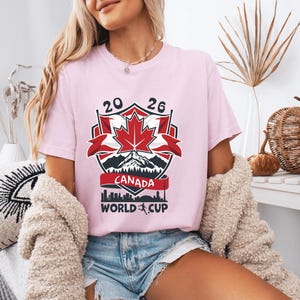 Peut inclure: T-shirt rose clair avec un motif graphique pour la Coupe du Monde Canada 2026. Le motif comprend une feuille d'&eacute;rable rouge, des montagnes et le texte "CANADA WORLD CUP".
