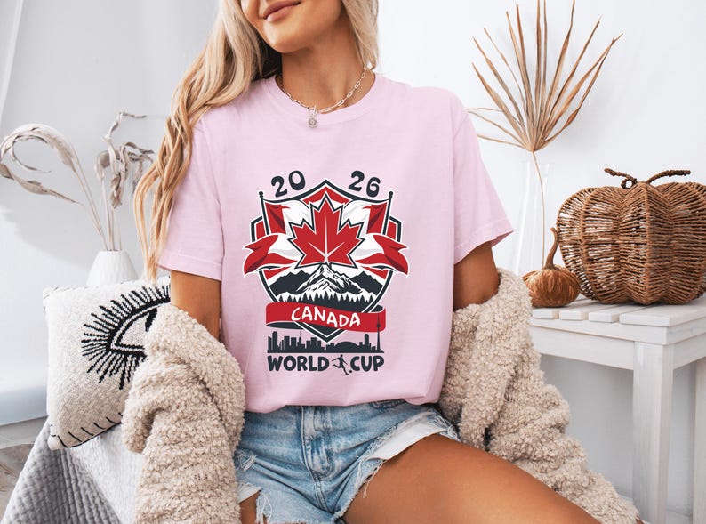 Peut inclure: T-shirt rose clair avec un motif graphique pour la Coupe du Monde Canada 2026. Le motif comprend une feuille d'&eacute;rable rouge, des montagnes et le texte "CANADA WORLD CUP".