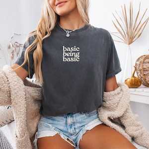 Puede incluir: Camiseta gris oscuro con las palabras "basic being basic" en letras blancas. La camiseta de manga corta tiene cuello redondo. La modelo lleva pantalones cortos vaqueros y una rebeca color crema.