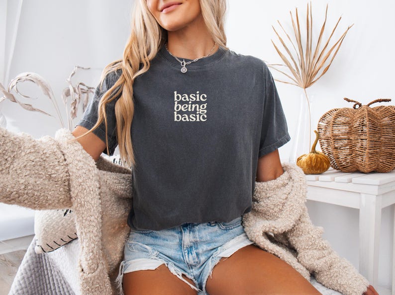 Puede incluir: Camiseta gris oscuro con las palabras "basic being basic" en letras blancas. La camiseta de manga corta tiene cuello redondo. La modelo lleva pantalones cortos vaqueros y una rebeca color crema.