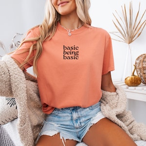 Puede incluir: Camiseta color coral con el texto "basic being basic" en negro. Se combina con shorts vaqueros y una rebeca crema y difusa. El conjunto transmite un estilo relajado y c&oacute;modo.