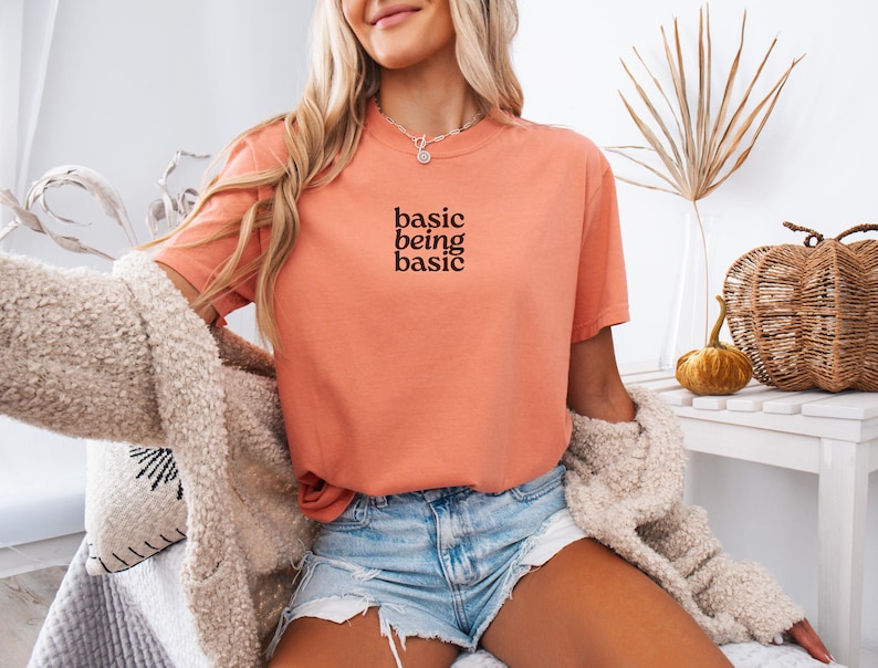Puede incluir: Camiseta color coral con el texto "basic being basic" en negro. Se combina con shorts vaqueros y una rebeca crema y difusa. El conjunto transmite un estilo relajado y c&oacute;modo.