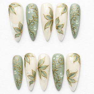 May include: A set of ten long, almond-shaped artificial nails. The nails feature a combination of white and pale green bases, adorned with gold floral and vine designs. Some nails have small, raised white dots.