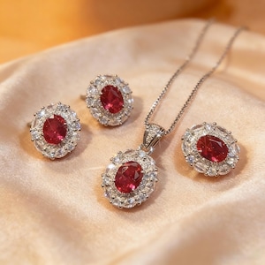 Puede incluir: Un conjunto de joyas que incluye un collar, un anillo y pendientes. Cada pieza tiene una gema roja ovalada central rodeada de piedras claras más pequeñas. El conjunto se exhibe sobre una superficie satinada.