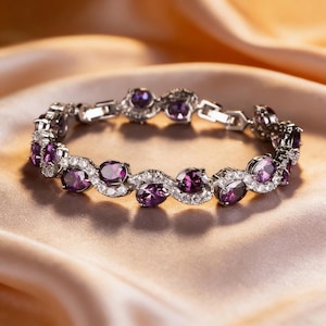 May include: A silver bracelet with oval-cut purple gemstones and small white accent stones. The bracelet is set on a satin surface, creating a luxurious feel. The gemstones are a deep, rich purple color.