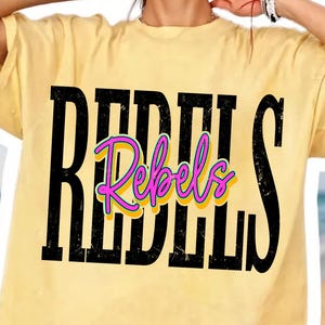 May include: A pale yellow short-sleeve t-shirt featuring the word REBELS in large, distressed black capital letters. Overlaid in the center is the word Rebels written in a bright pink cursive script with a yellow outline and a thin black border.