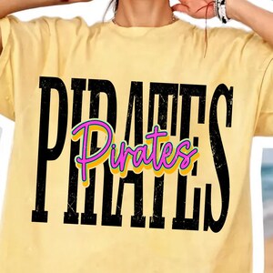 May include: A pale yellow short-sleeve t-shirt featuring the word PIRATES in large, distressed black capital letters. Overlaid in the center is the word Pirates in a bright pink script font with a yellow outline, creating a layered effect.