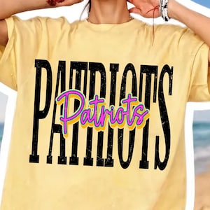 May include: A pale yellow short-sleeve t-shirt featuring the word PATRIOTS in large, distressed black capital letters. Overlaid in the center is the word Patriots in a bright pink cursive font with a yellow outline and a thin purple border.