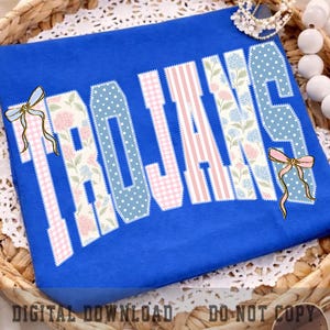 May include: A royal blue t-shirt featuring the word TROJANS in large block letters. The letters are filled with alternating patterns of pink and white stripes, blue and white polka dots, and floral prints. Two light blue and pink bows are attached.