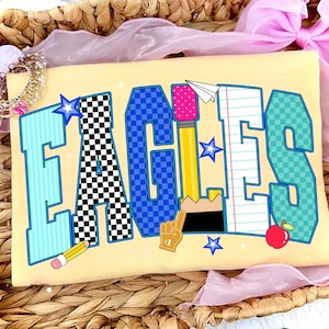 May include: A light yellow t-shirt featuring the word EAGLES in large block letters. Each letter has a unique pattern: light blue stripes, black and white checkers, blue checkers, a yellow pencil, notebook paper, and teal checkers.