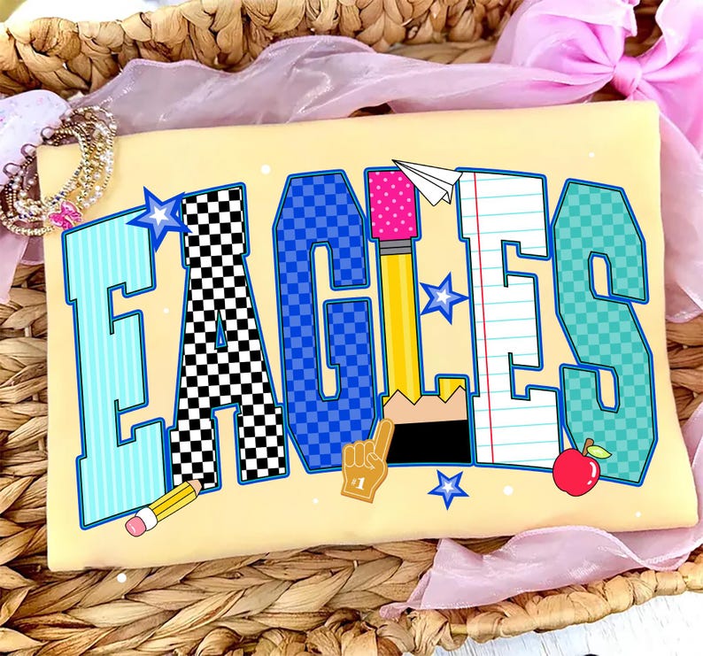 May include: A light yellow t-shirt featuring the word EAGLES in large block letters. Each letter has a unique pattern: light blue stripes, black and white checkers, blue checkers, a yellow pencil, notebook paper, and teal checkers.