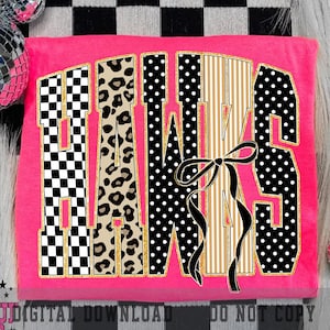 May include: A bright pink t-shirt featuring the word HAWKS in large block letters. Each letter has a unique pattern including black and white checkers, leopard print, black with white polka dots, and beige stripes, all outlined in gold glitter.