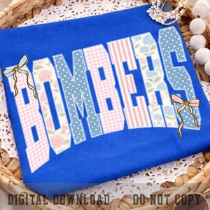 May include: A royal blue t-shirt featuring the word BOMBERS in large block letters. Each letter has a unique pattern including pink gingham, blue polka dots, pink and white stripes, and floral prints. Two light blue and pink bows accent the text.