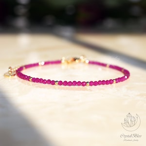 May include: A delicate bracelet featuring small, round, magenta-colored beads interspersed with gold-colored accents. The bracelet has a gold clasp and a small, round charm. The jewelry is displayed on a light-colored surface.
