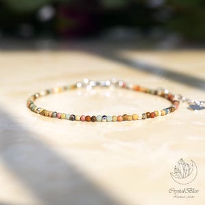 Puede incluir: Delicada pulsera de cuentas con pequeñas piedras preciosas de colores en tonos marrones, verdes, naranjas y rosas. La pulsera tiene un cierre plateado y un pequeño dije de corazón. La joya se exhibe sobre una superficie clara.