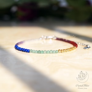May include: A colourful beaded bracelet featuring a spectrum of colours from blue to red. The bracelet has a silver clasp and a small charm. The background is blurred, with a logo in the bottom right corner.