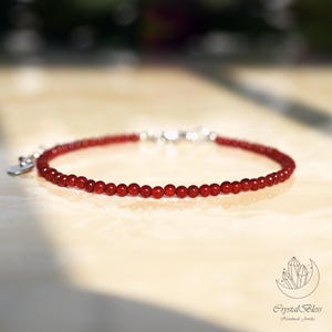 May include: A delicate bracelet featuring small, round, red beads strung together. The bracelet has a silver clasp and a small heart-shaped charm. The jewelry is displayed on a light-coloured surface.