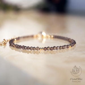 May include: A delicate bracelet featuring small, faceted, smoky quartz beads strung together with gold-colored accents. The bracelet has a small heart-shaped charm and a clear, round bead. The jewelry is displayed on a reflective surface.