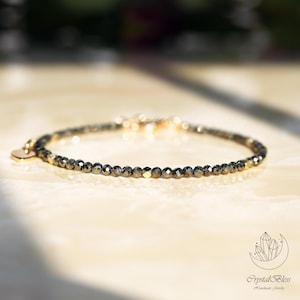 Puede incluir: Una pulsera delicada con pequeñas cuentas facetadas de color gris oscuro. Un pequeño dije en forma de corazón dorado cuelga de la pulsera. La pulsera se exhibe sobre una superficie clara, con iluminación suave.