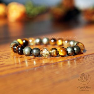 May include: A beaded bracelet featuring a mix of polished gemstones in shades of gold, brown, and silver. The bracelet is displayed on a wooden surface, with the "Crystal Bliss Handmade Jewelry" logo in the bottom right corner.