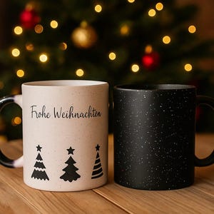 Könnte beinhalten: Zwei Keramikbecher mit schwarzen Griffen. Einer ist cremefarben mit dem Text "Frohe Weihnachten" und schwarzen Tannenbaum-Designs. Der andere ist schwarz mit weißen Sprenkeln. Der Hintergrund ist ein Weihnachtsbaum mit Lichtern.