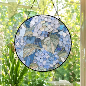 May include: Round stained glass window art featuring a hydrangea design. The artwork showcases blue and white flowers with green leaves, set against a mosaic of blue glass. A black metal frame and chain complete the piece.