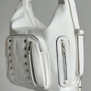 Può includere: Borsa a tracolla bianca con finitura strutturata. La borsa presenta più tasche con cerniera e dettagli di rivetti color argento. È attaccata una lunga tracolla regolabile. La borsa è realizzata in materiale resistente.