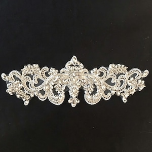 May include: A silver-toned embroidered appliqué featuring intricate scrollwork and floral patterns. The design is heavily embellished with clear, faceted crystals and beads, creating a symmetrical, elongated shape with a pointed centre element.