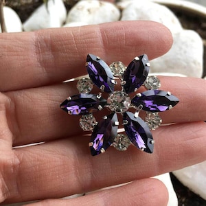 May include: A flower-shaped brooch with a silver setting. The brooch features six deep purple marquise-cut crystals arranged as petals, surrounding a central clear crystal. Smaller clear crystals are interspersed between the petals.