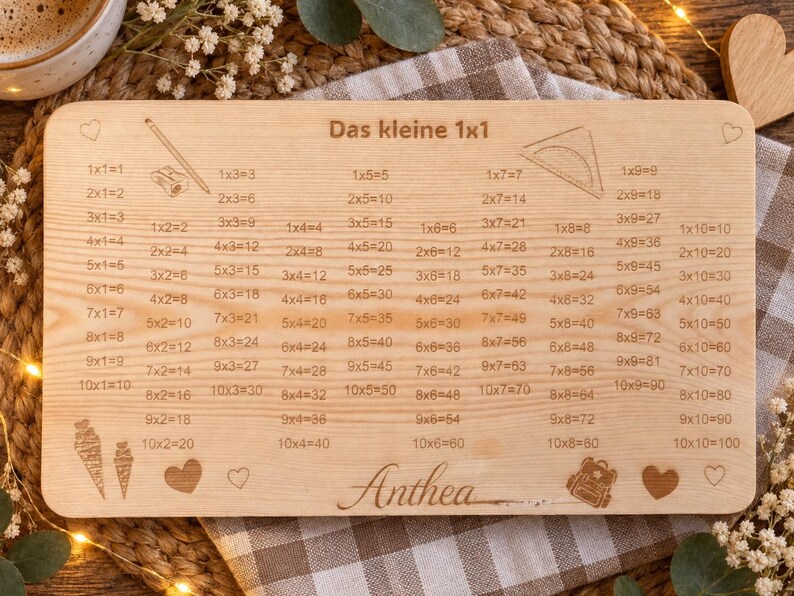 May include: A rectangular light-colored wooden board featuring the engraved text Das kleine 1x1 and a comprehensive multiplication table from 1x1 to 10x10. The board includes engraved illustrations of a pencil, a protractor, ice cream cones, a backpack, and hearts, with the name Anthea engraved at the bottom center.