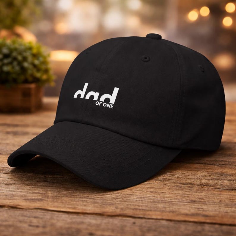 Op de afbeelding: Zwarte baseballcap met het woord "dad" in witte, gestileerde letters, boven de woorden "of one". De cap heeft een gebogen klep en een stoffen bandsluiting. De hoed ligt op een houten oppervlak.