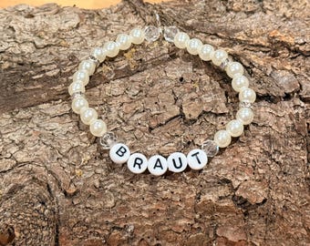 May include: A pearl bead bracelet with the word "BRAUT" spelled out in white letter beads. The bracelet features clear crystal accents and is displayed on a textured wooden surface.