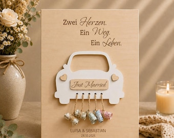May include: A rectangular wooden plaque featuring the German text "Zwei Herzen. Ein Weg. Ein Leben." and a white wooden car cutout with "Just Married" text. Five rolled banknotes hang from the car by twine. Below, "LUISA & SEBASTIAN 24.10.2026" is engraved.