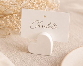 May include: A white heart-shaped card holder made of a smooth material, holding a rectangular white place card. The card features the name Charlotte in a dark script font with a small heart icon centered above the name in a light color.