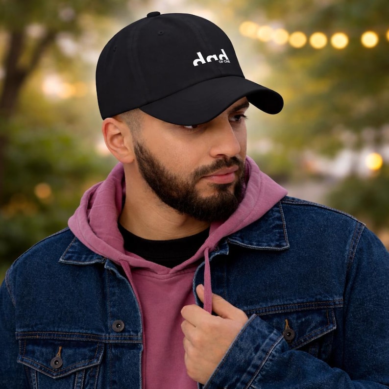 Op de afbeelding: Zwarte baseballcap met de witte tekst "DAD OF ONE". De cap heeft een gebogen klep en is gemaakt van een zacht materiaal. De persoon op de foto draagt een roze hoodie en een spijkerjas.