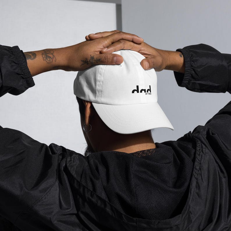 Op de afbeelding: Een witte baseball cap met de zwarte tekst "dad of two". De cap heeft een gebogen klep en is gemaakt van een zacht materiaal. Gedragen door een persoon in een zwarte jas.
